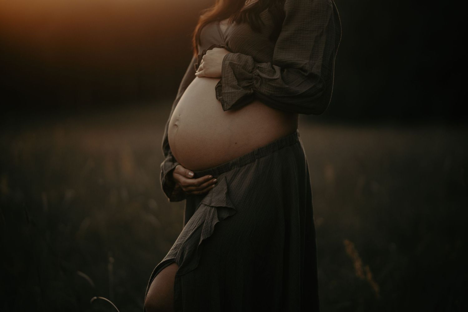A pregnant woman in a floral dress stands in a golden field at sunset.
