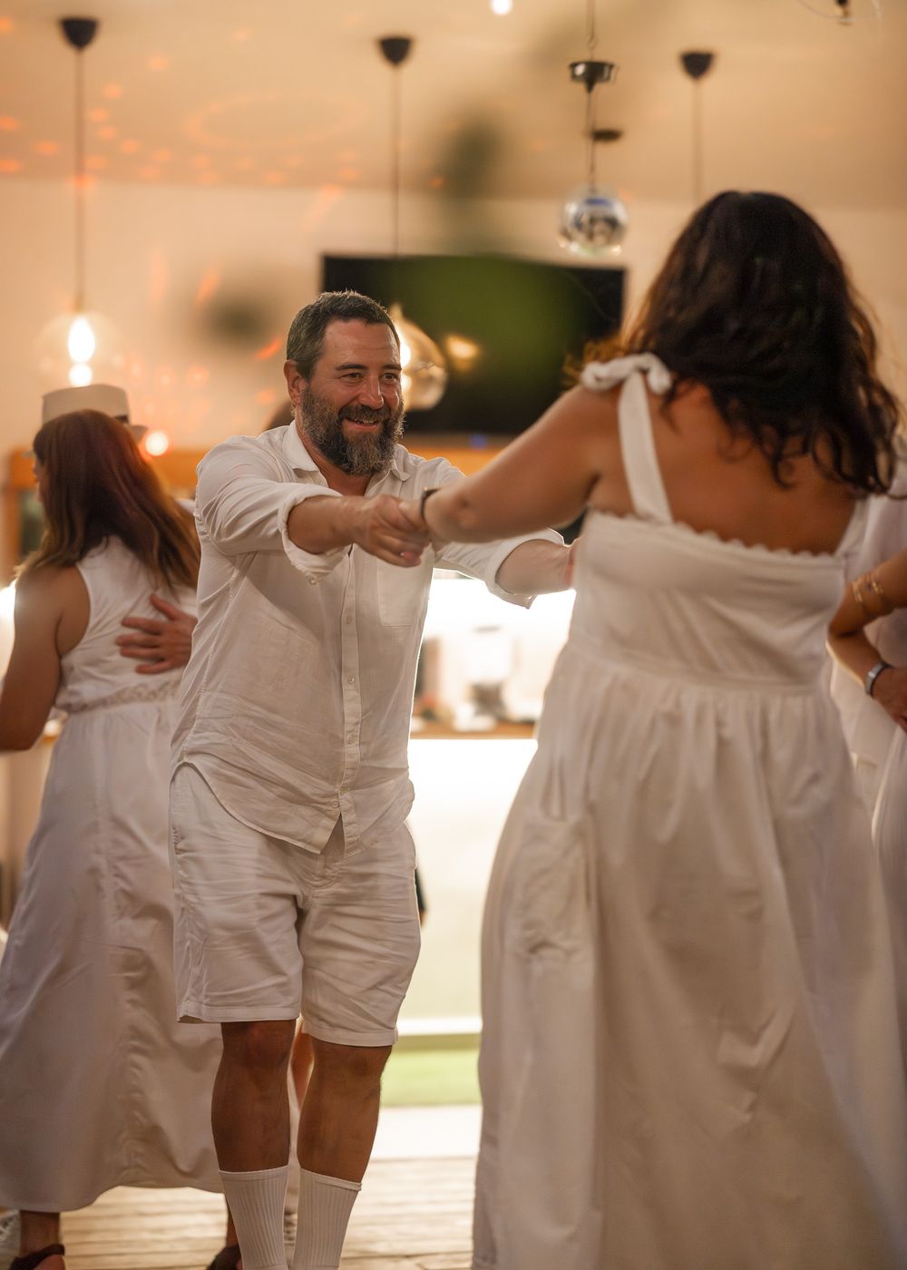 People in white clothing dance together at an indoor celebration with warm lighting and pendant lamps overhead.