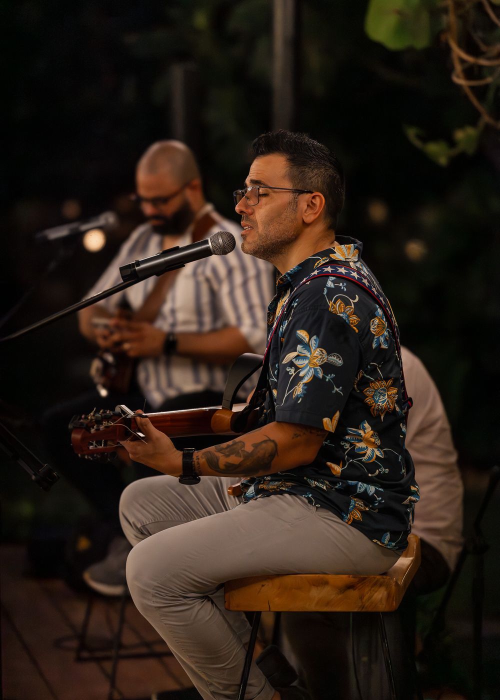 Musician in a floral shirt performs on stage with acoustic guitar during an evening outdoor concert.