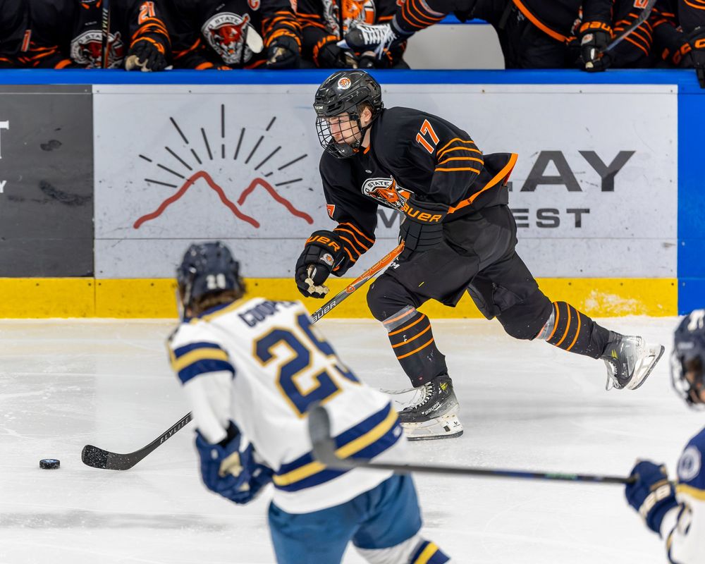 Hockey player in black and orange uniform skates with the puck during an intense game against opponent in blue and white.