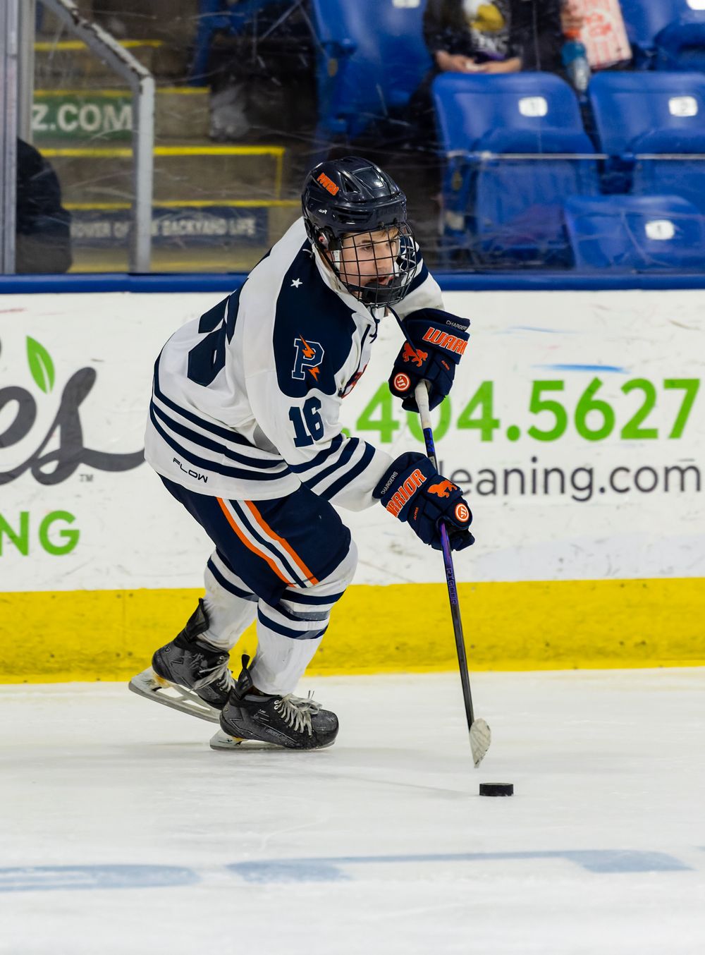 A hockey player wearing a white and orange jersey is skating on an ice rink with a hockey stick in hand.