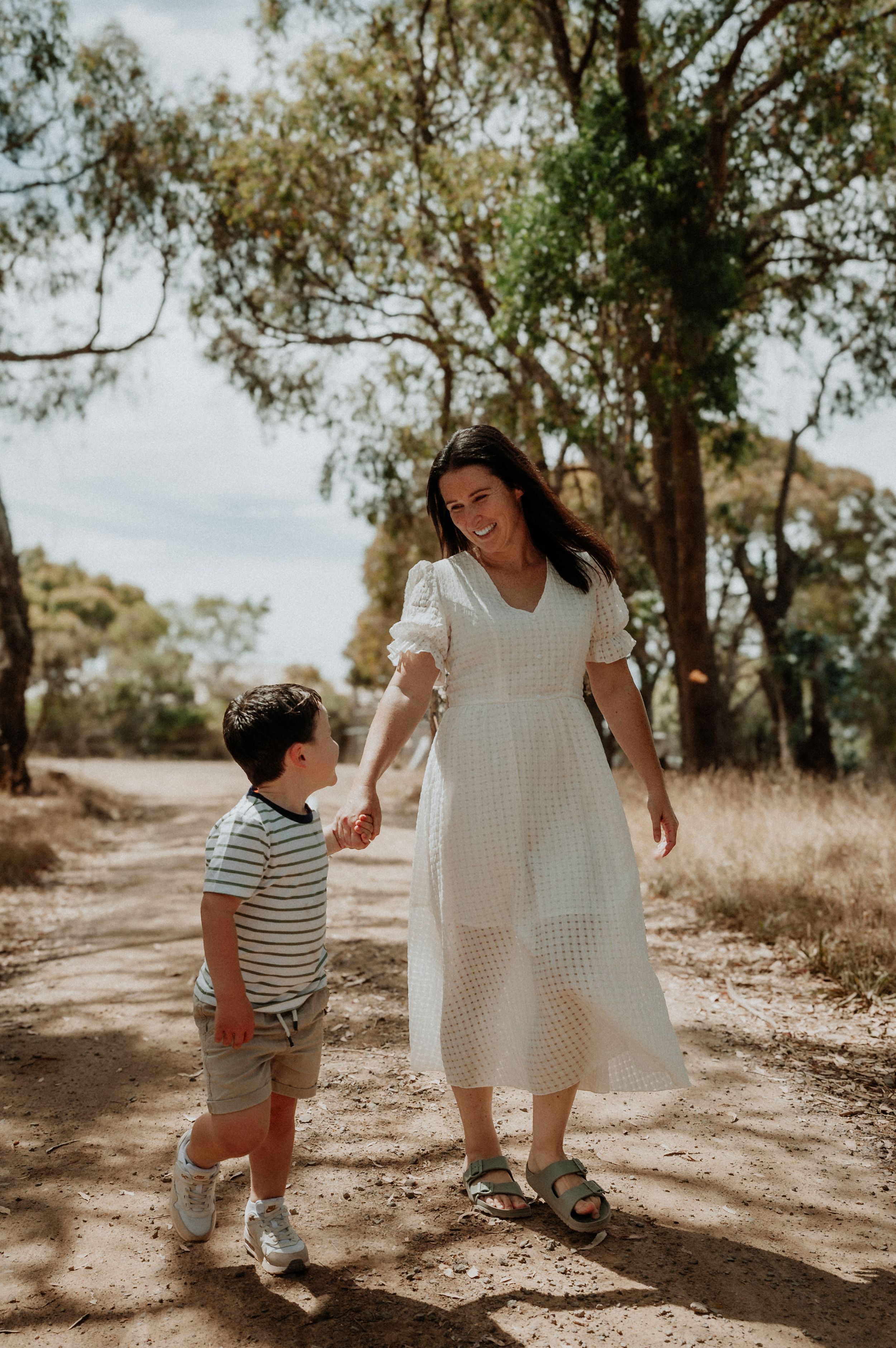 A mother and child walk hand in hand down a sunlit dirt path surrounded by eucalyptus trees.
Altona Family Photographer Melbourne