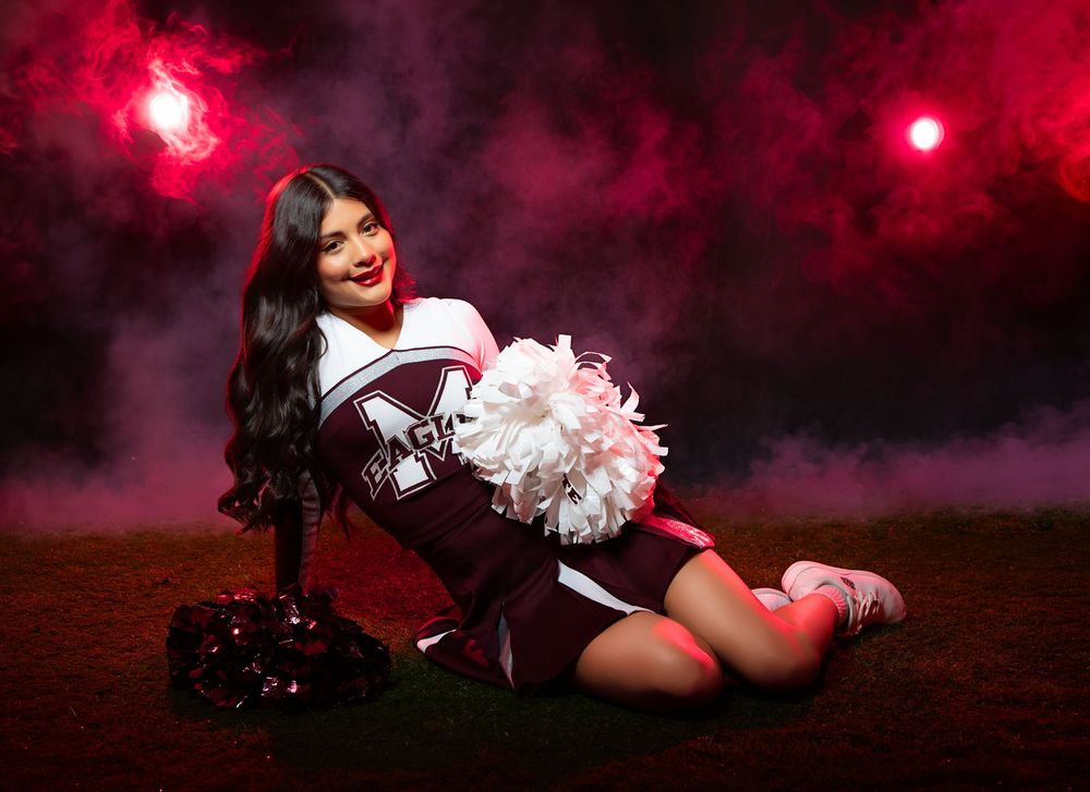 Cheerleader in maroon uniform poses with pom-poms against dramatic red smoky background on field.