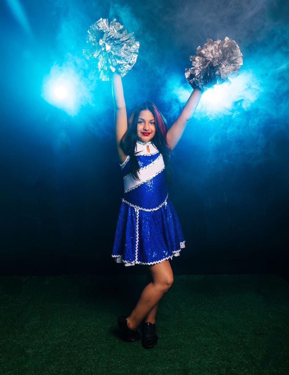 Cheerleader in blue sequin uniform holding silver pom-poms with dramatic blue smoke lighting backdrop.