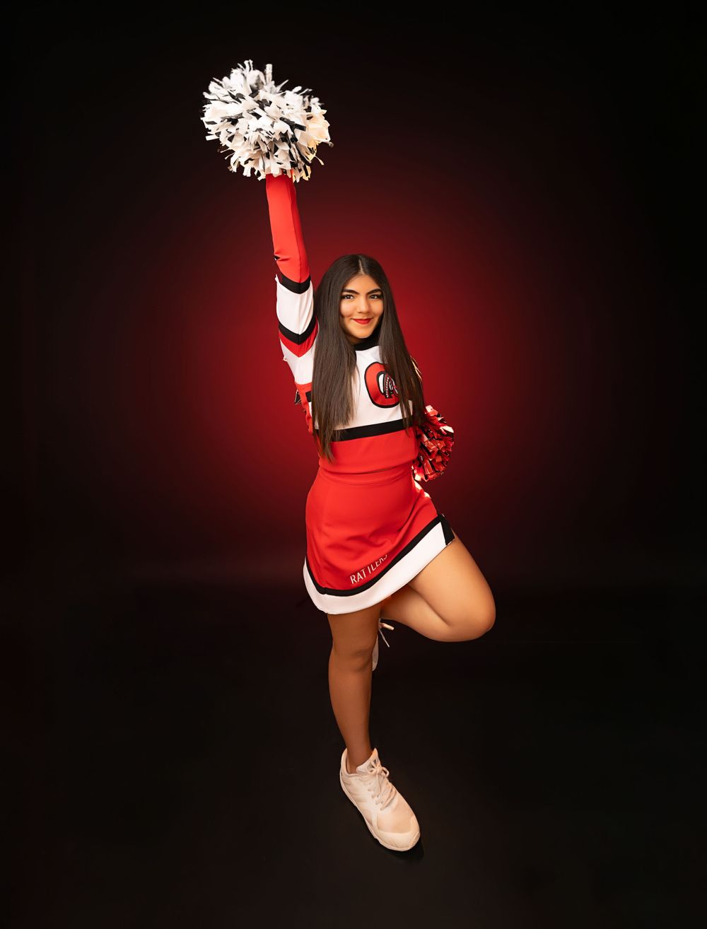 Cheerleader in dynamic pose holding white pom pom overhead against dark background.
