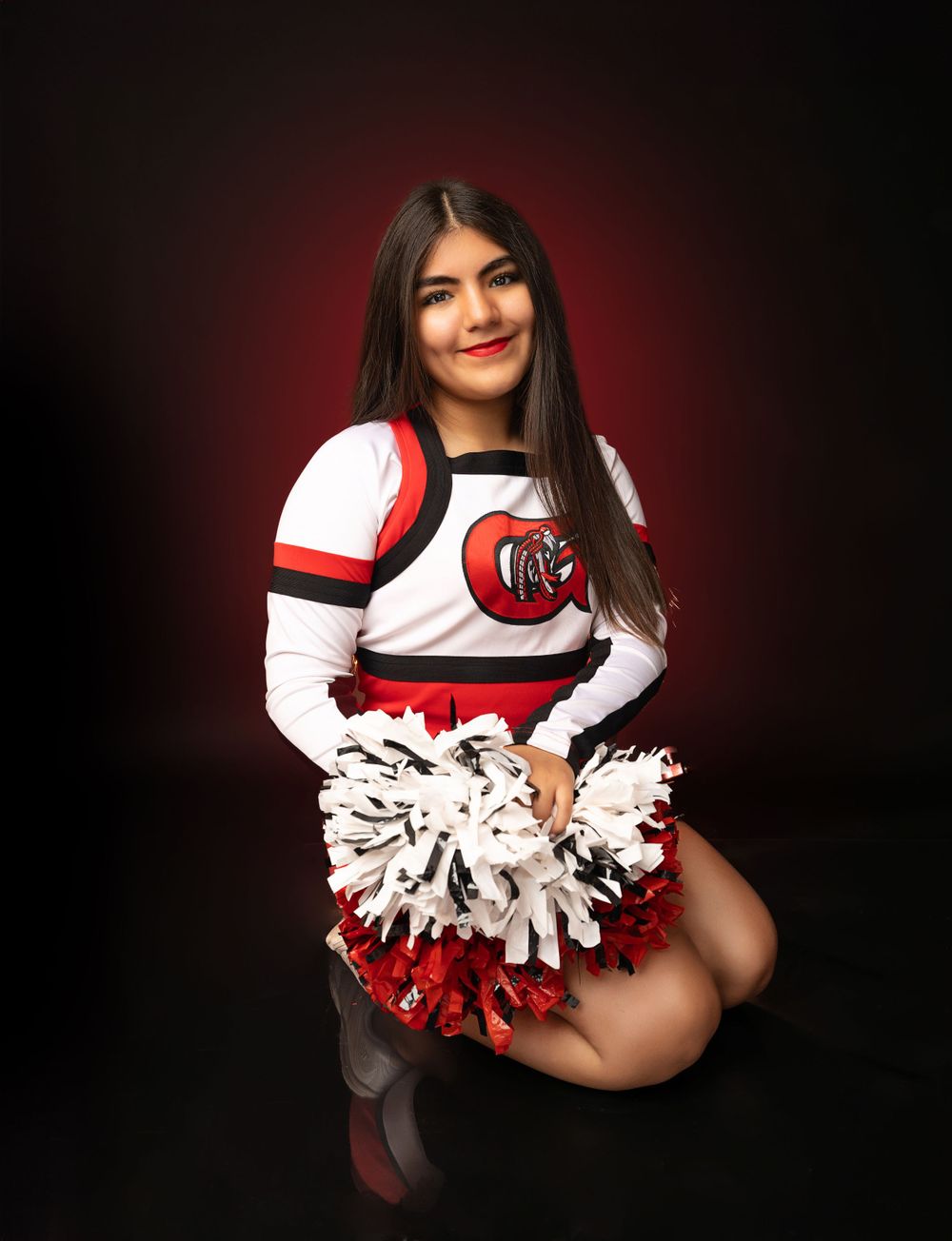 Cheerleader kneeling with red and white pom poms in studio portrait with red background glow.