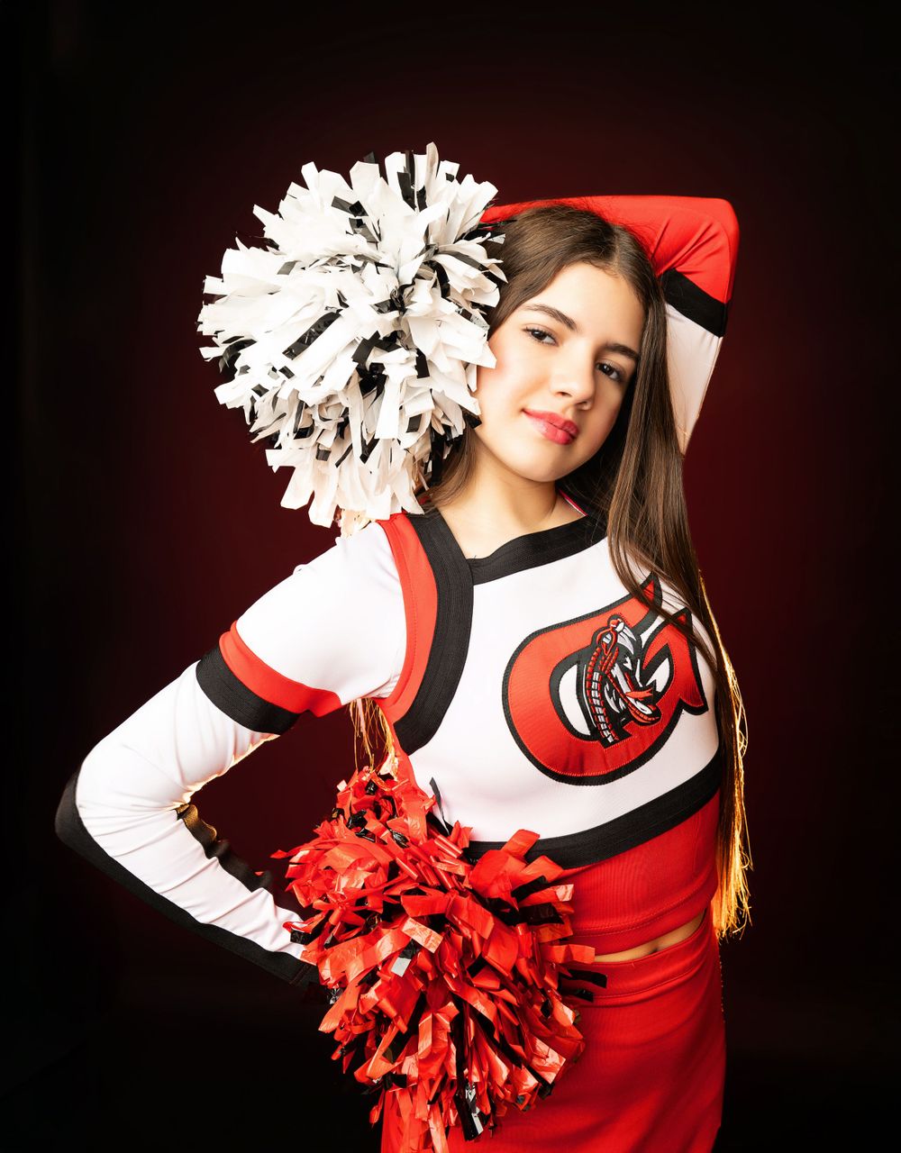 Cheerleader strikes dynamic pose with red and white pom poms against dark studio backdrop.