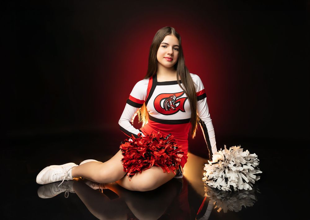 Cheerleader sitting with red and white pom poms in studio with red backlight effect.