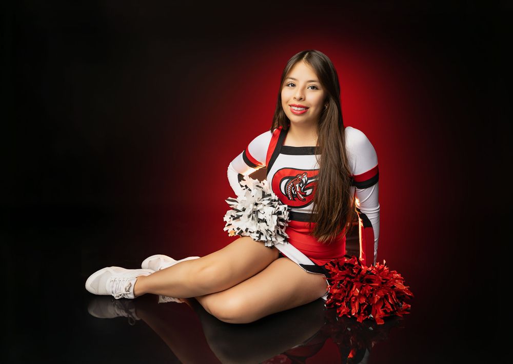 Cheerleader poses with red and white pom poms on dark floor with dramatic red lighting.
