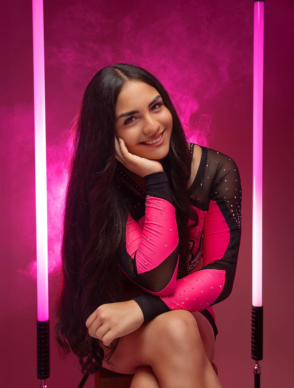 A person in a black and hot pink outfit poses against a pink-lit backdrop with vertical light bars.