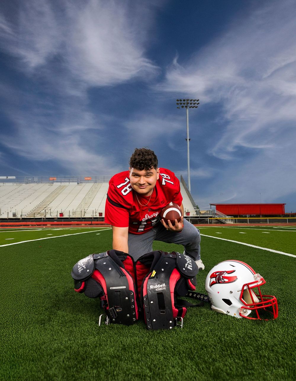 High school football player in red uniform poses with helmet and shoulder pads on green field under blue sky.