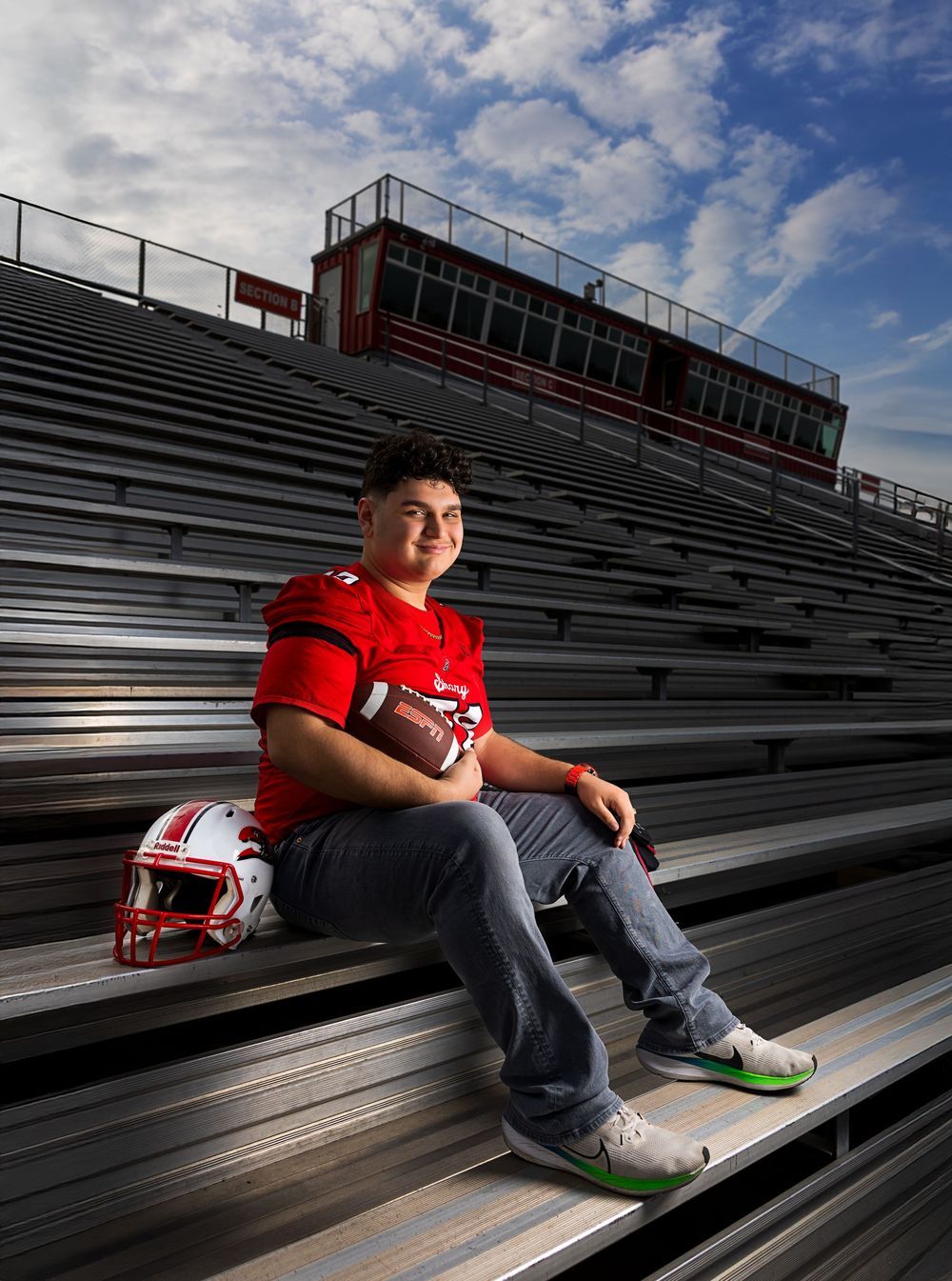 Young football player in red jersey sits on stadium bleachers holding helmet, posing for senior portrait.