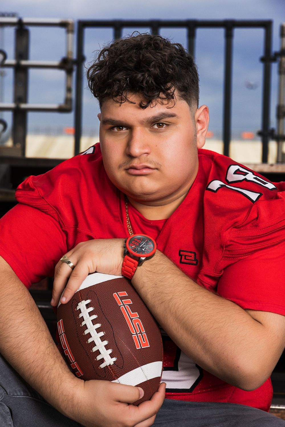 Young football player in red jersey holding a football, wearing a red watch, posed for a sports portrait photo.