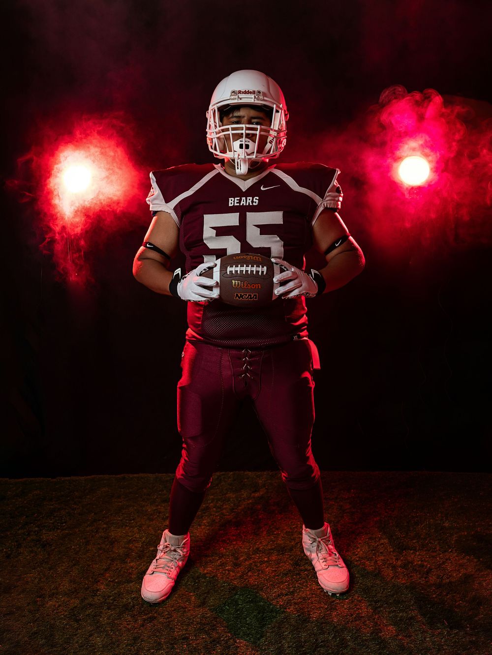 Football player #55 in maroon Bears uniform poses with ball under dramatic red smoke lights.