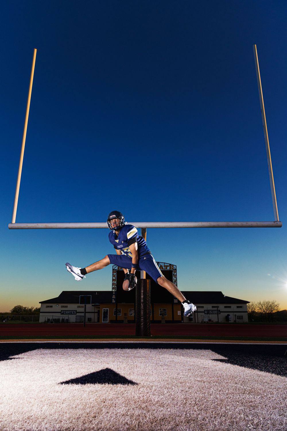 Football player in blue uniform jumps in front of goal posts at dusk on a lit field.