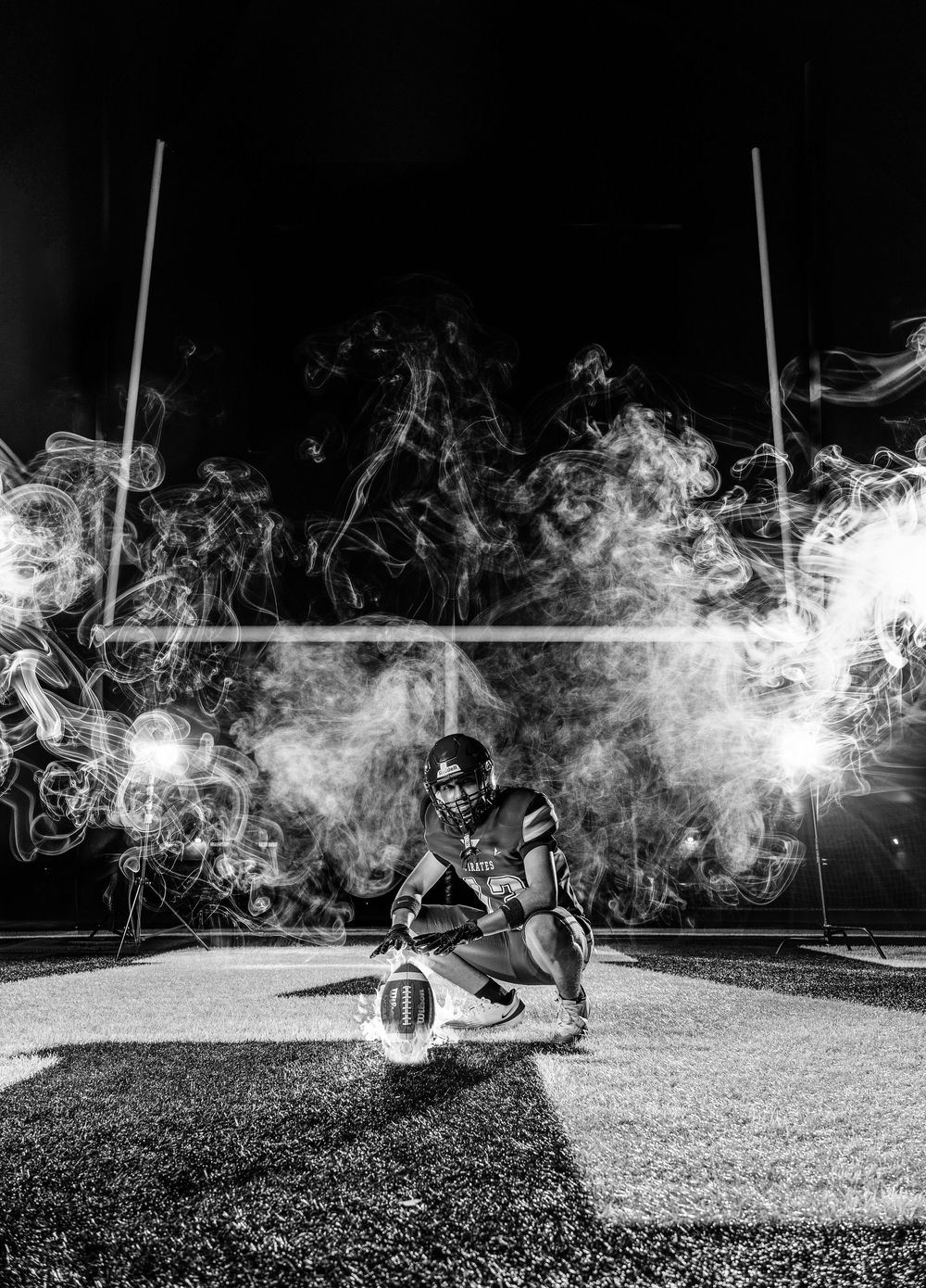 Black and white photo of a football player crouching on field at night surrounded by dramatic smoke and bright lights.
