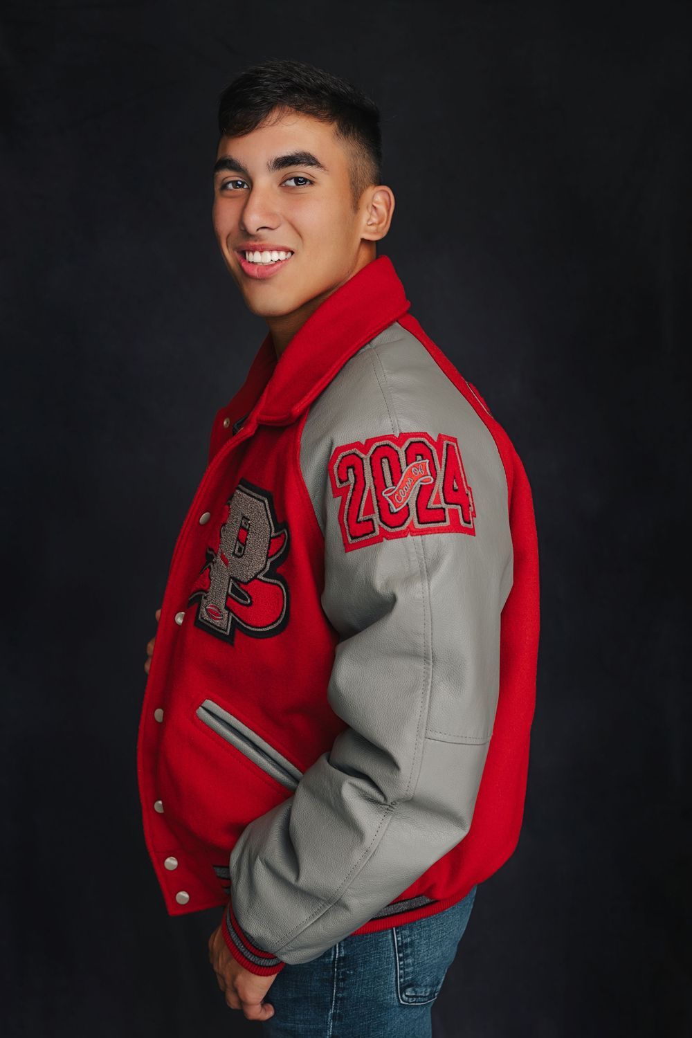 Smiling young man in red and gray 2024 varsity letterman jacket posing for a senior portrait against dark background.