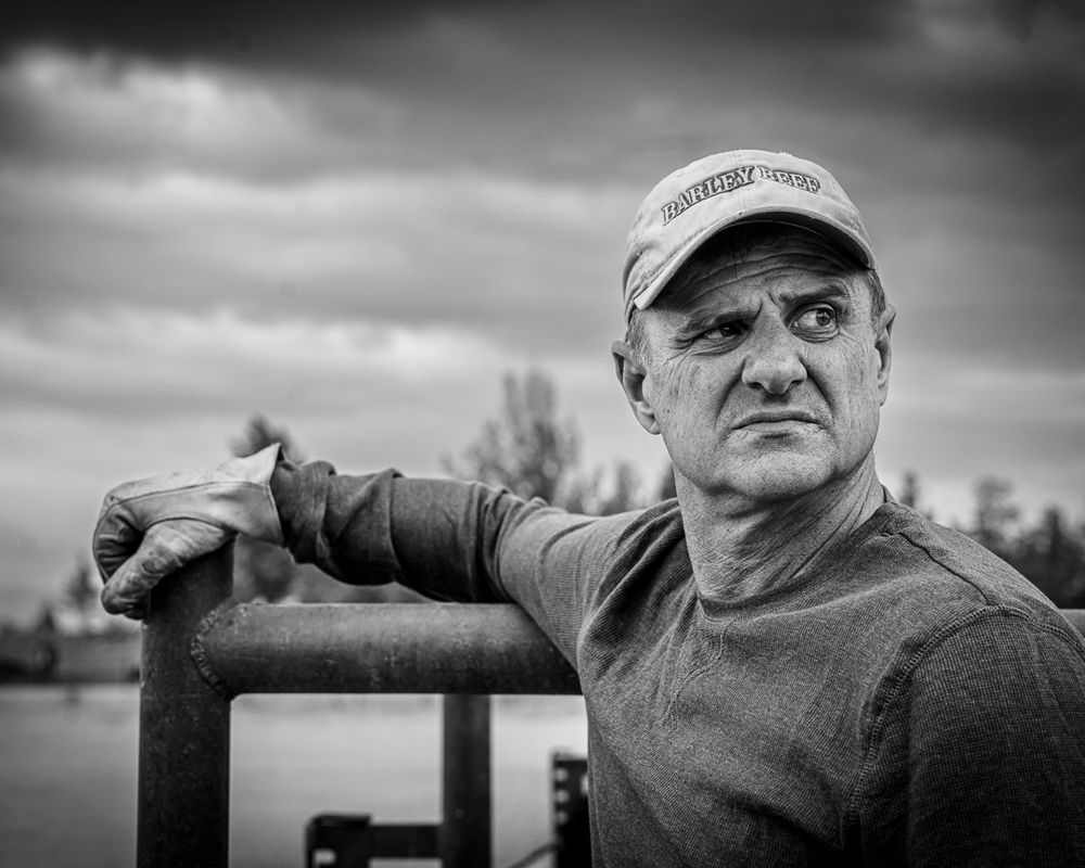 Black and white portrait of person wearing a baseball cap leaning on metal railing against cloudy sky backdrop.