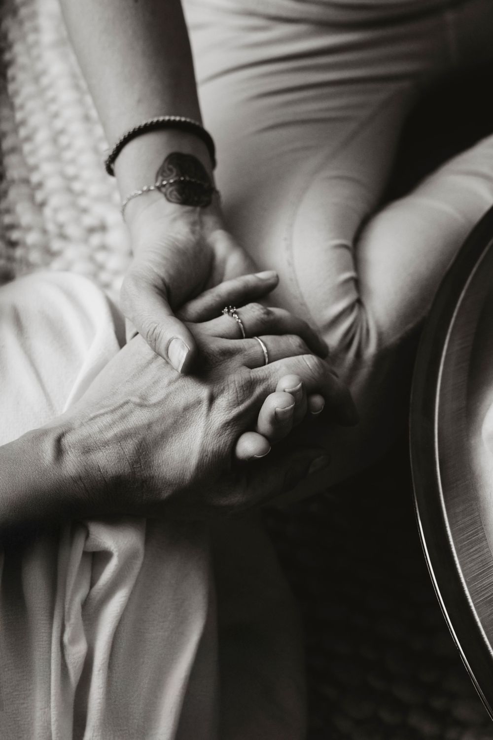 A black and white close-up photograph showing clasped hands with bracelets against white fabric background.