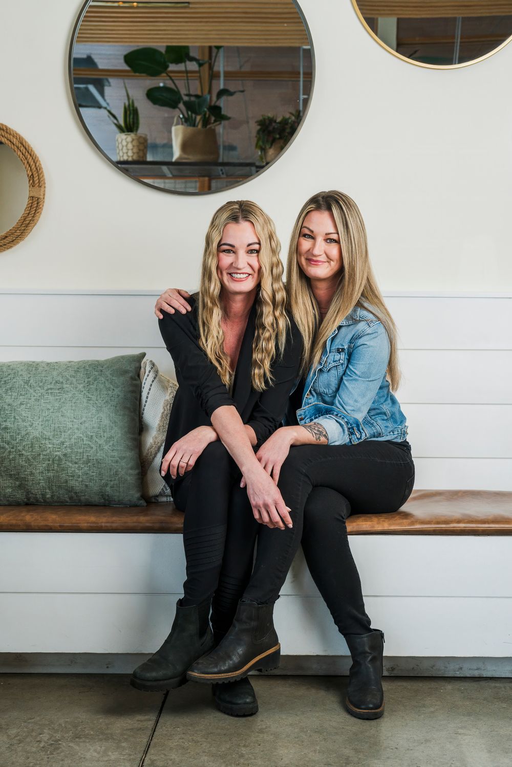 Two blonde women in casual outfits sitting together on a bench against a white wall with decorative round mirrors.