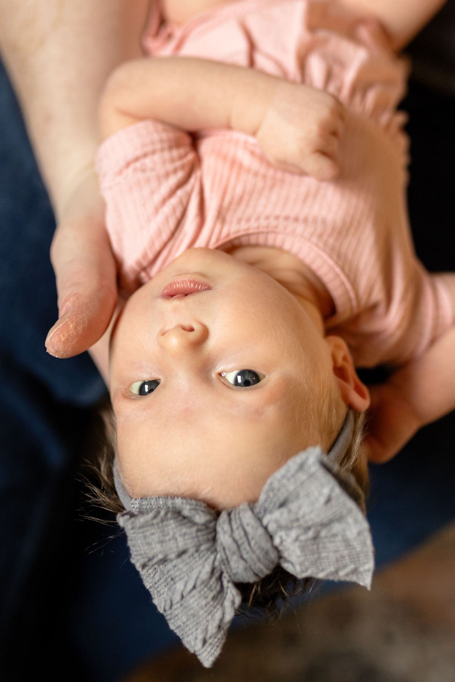 A baby wearing a pink shirt and large gray bow lies upside down while being held.
