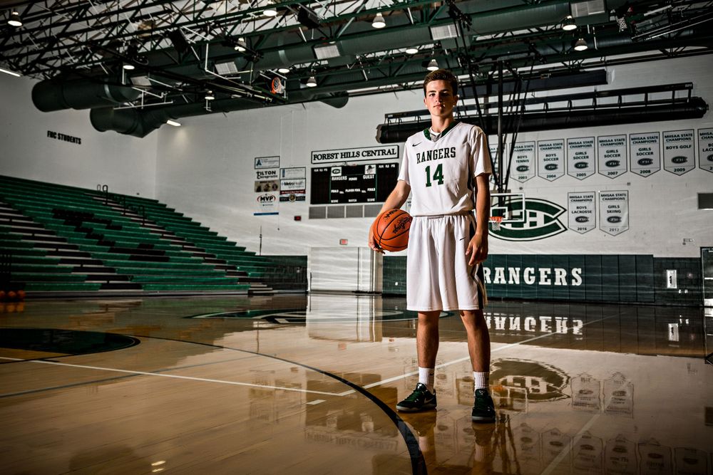 Basketball player in white uniform stands in empty school gymnasium with banners. Grand Rapids Photographer
