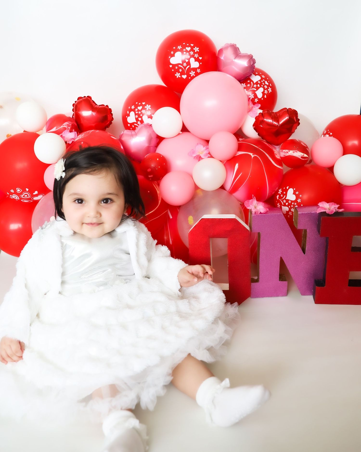 Baby in white dress sits among red and pink Valentine's Day balloon decorations.