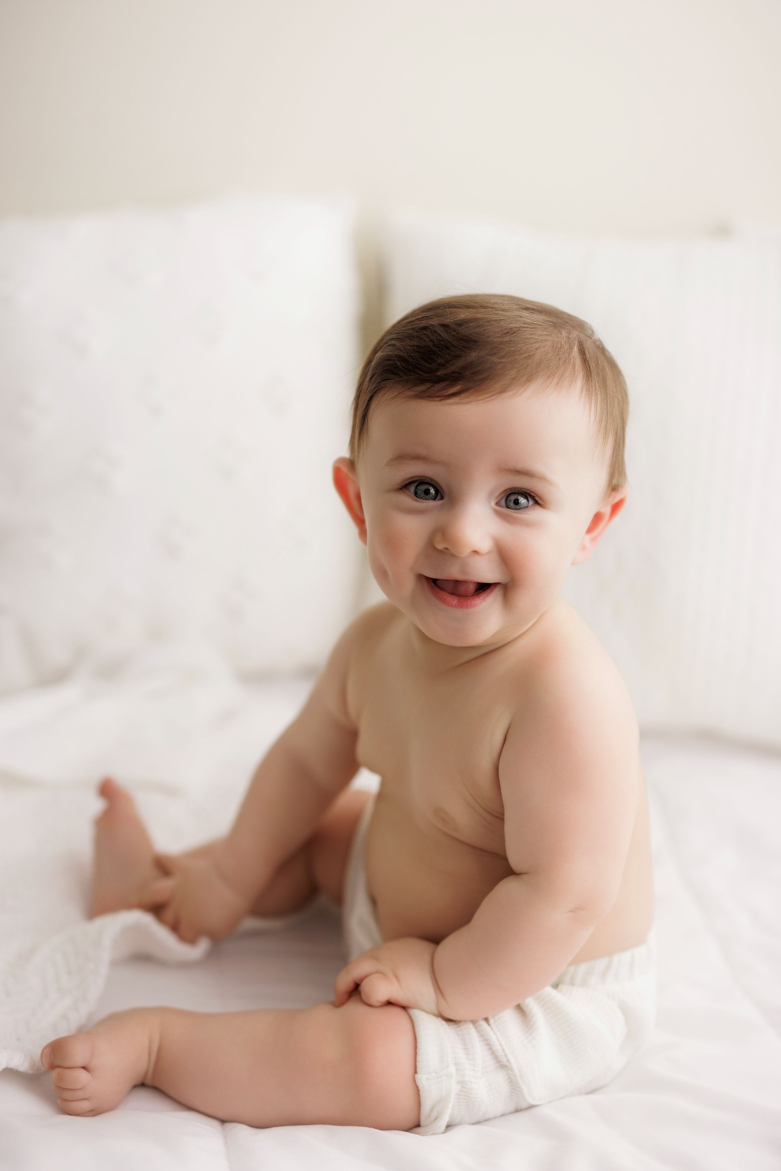 Happy baby wearing a white diaper sits on white bedding with a bright smile.