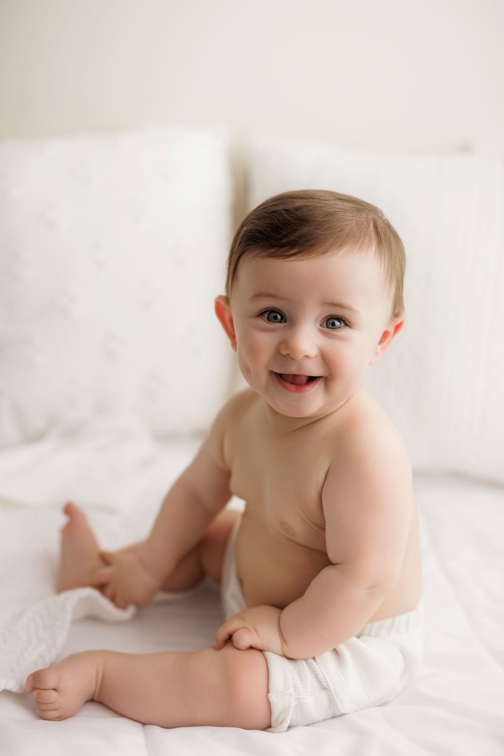 Happy baby wearing a white diaper sits on white bedding with a bright smile.