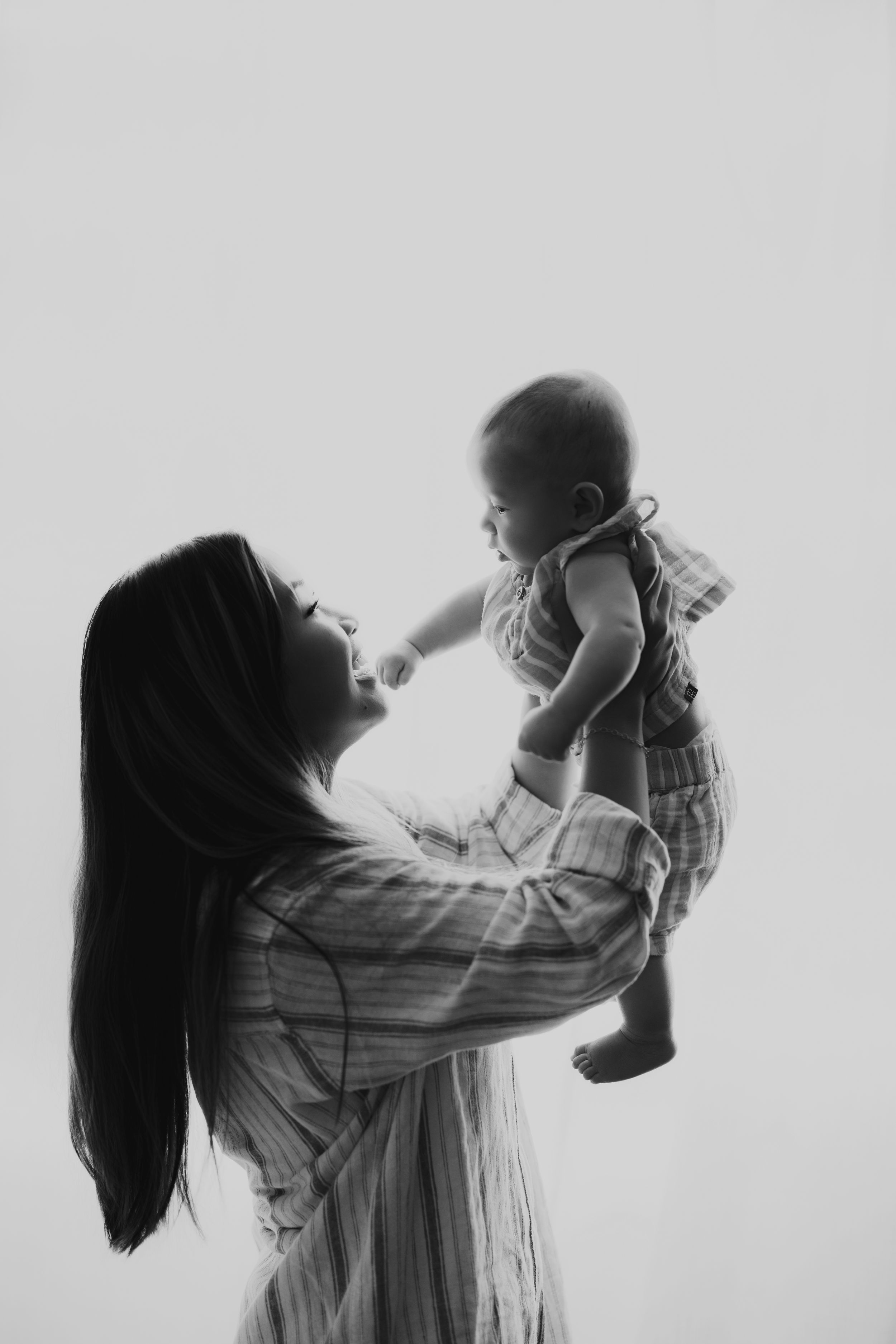 A parent in flowing dress lifts small child into the air against a bright background in black and white photograph.