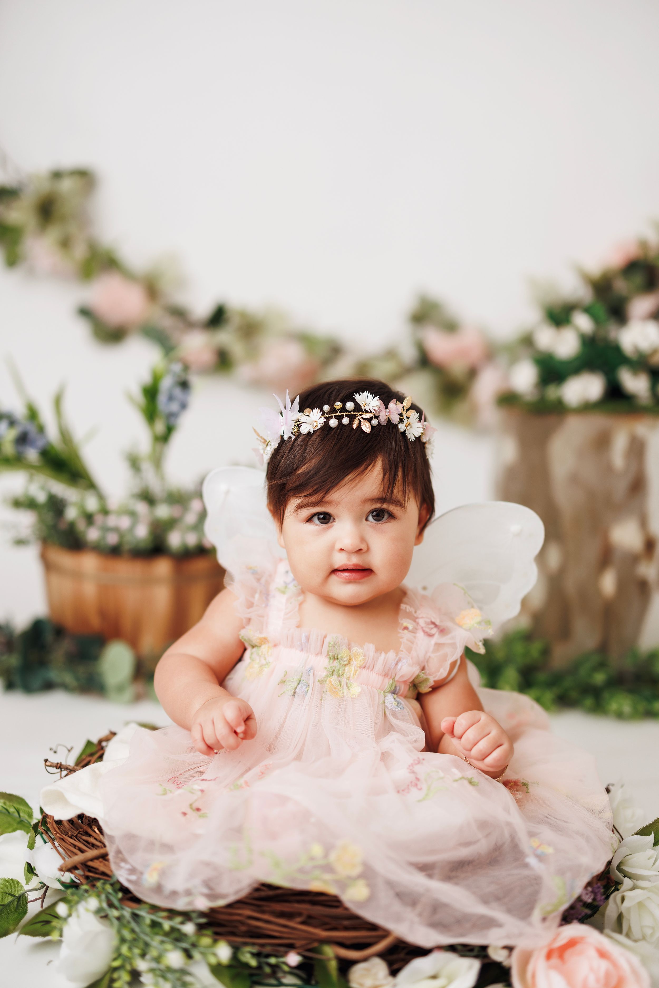 Baby girl in pink tulle dress and floral crown sitting in rustic basket with flowers.