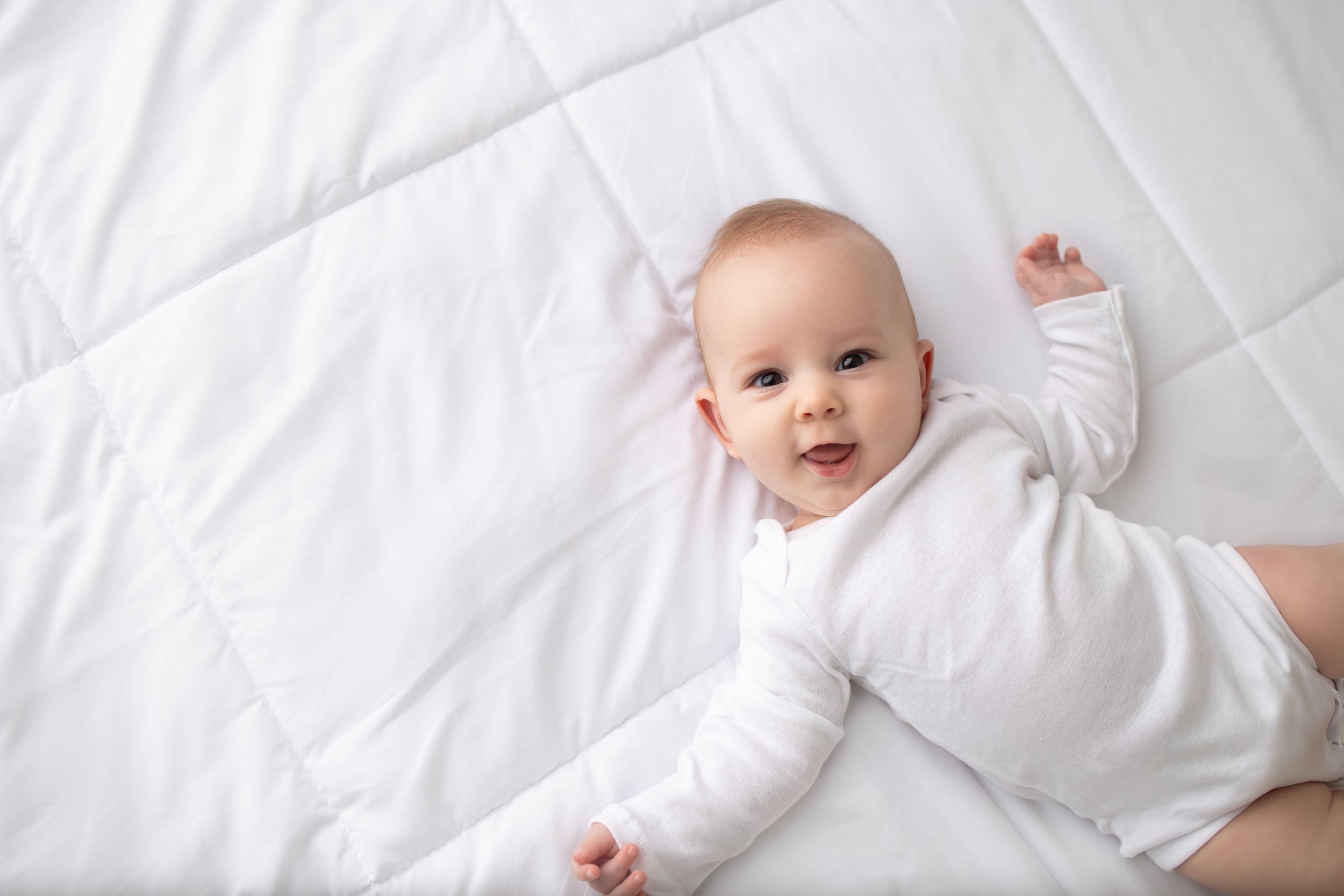 Baby wearing white onesie lying on soft white bedding.