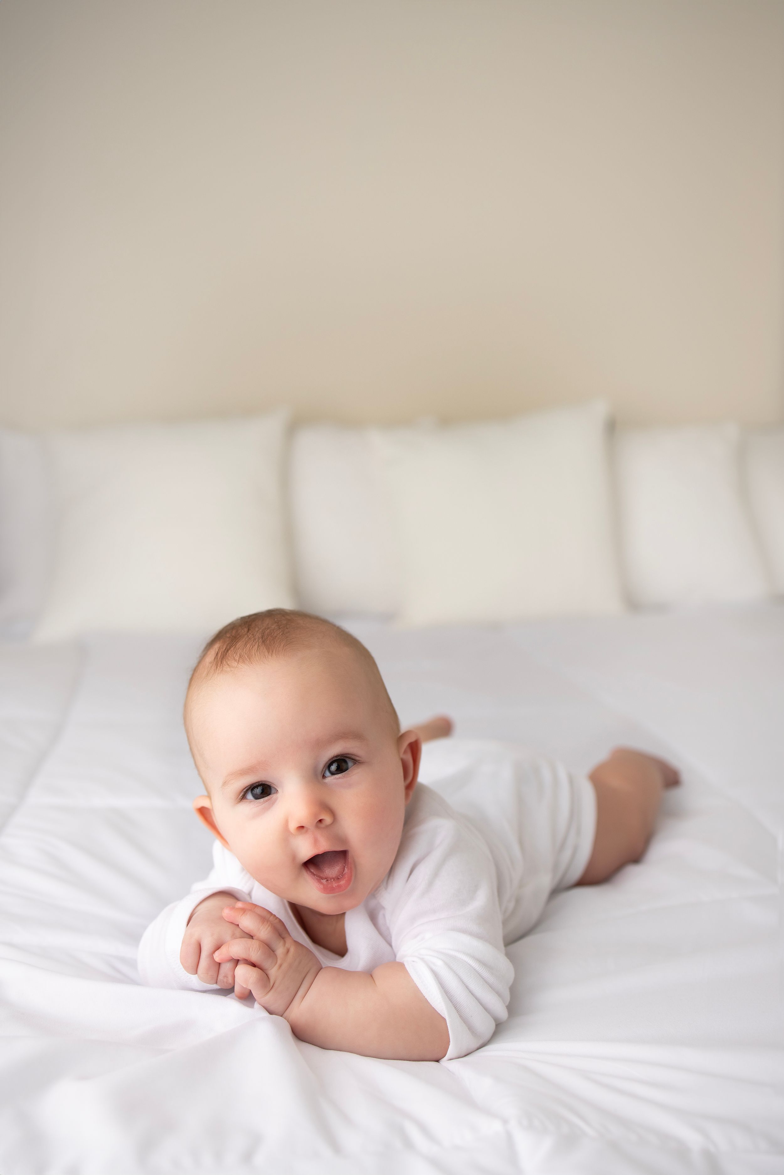 Happy baby in white onesie lying on bed with white pillows.