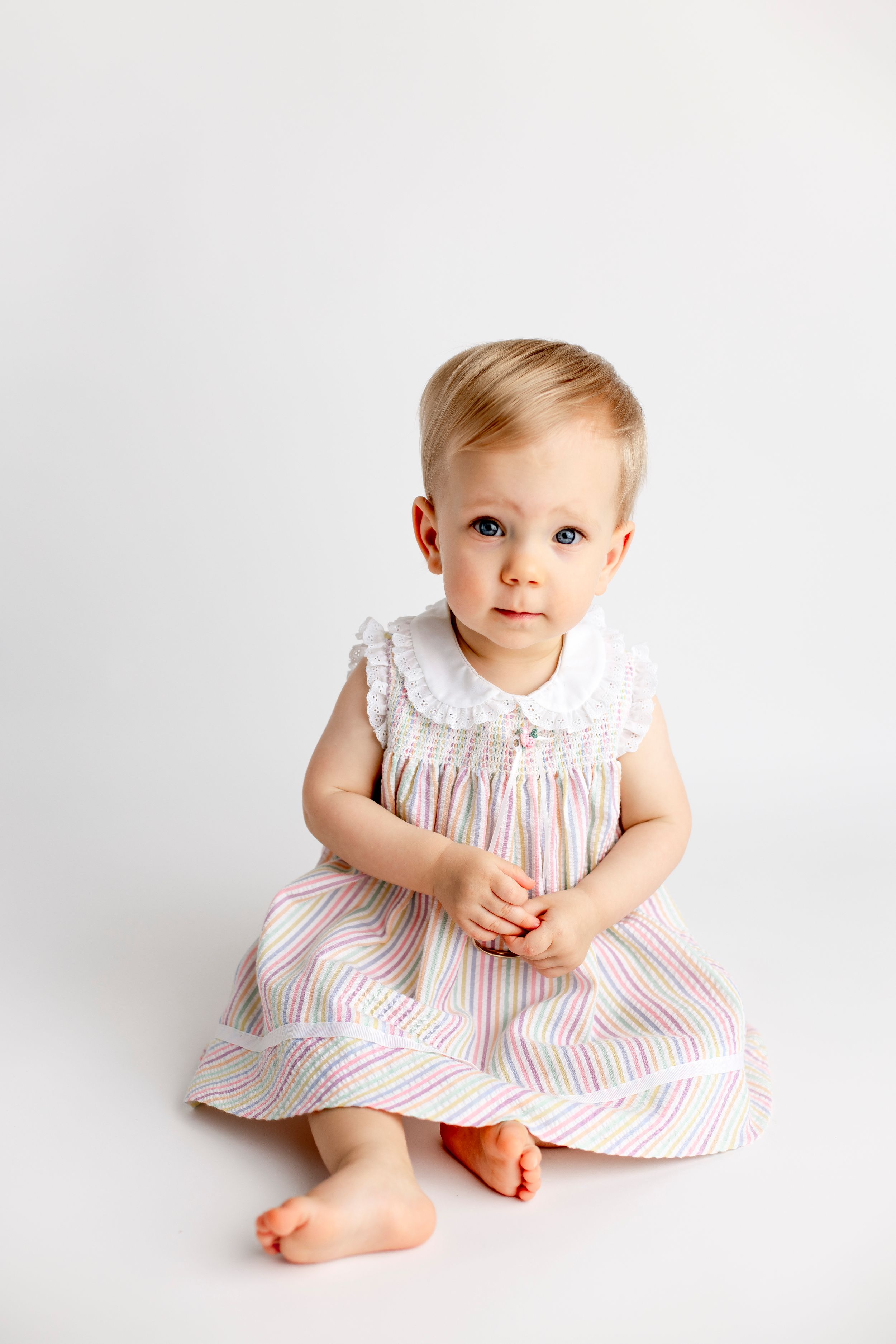 Baby in striped dress with lace collar sitting on white background.
