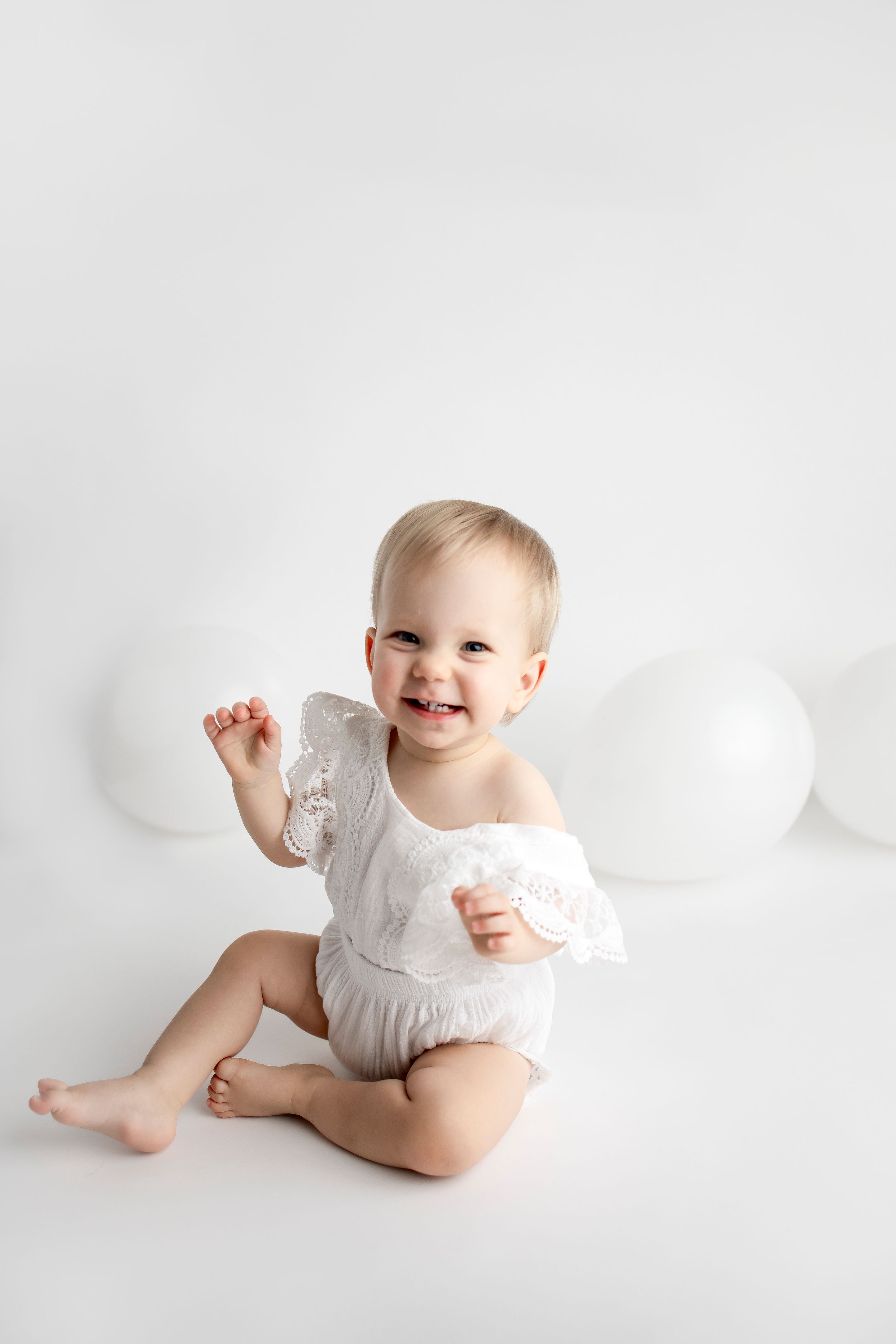 Baby in white ruffled outfit sitting on white background with balloons.
