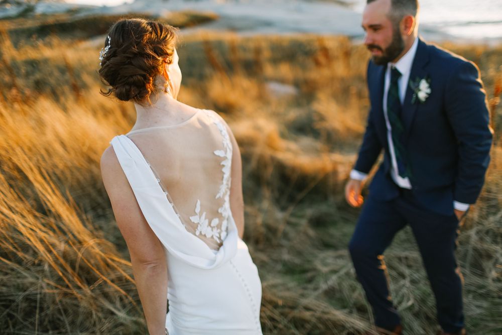 Back view of bride in lace gown with groom standing in tall golden grass during sunset Halifax, NS wedding photo session.