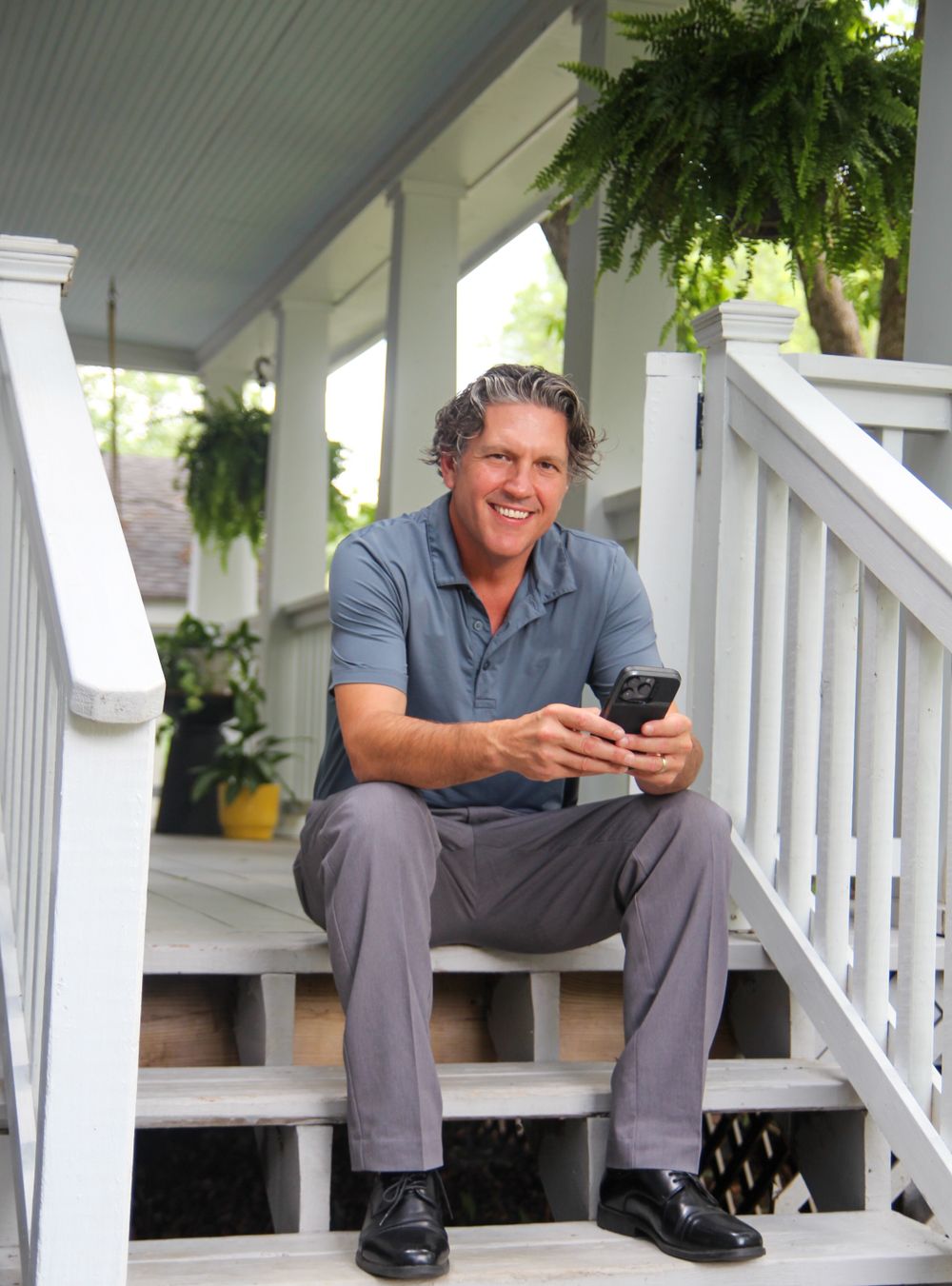 Person sits relaxed on white front porch steps holding a mobile phone, with hanging ferns decorating the space.