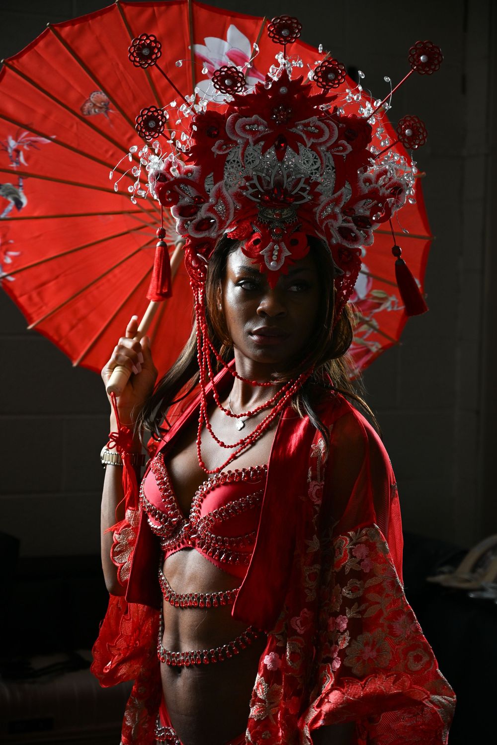 Stunning red Chinese-inspired costume with ornate headdress and parasol captured in dramatic dark lighting.