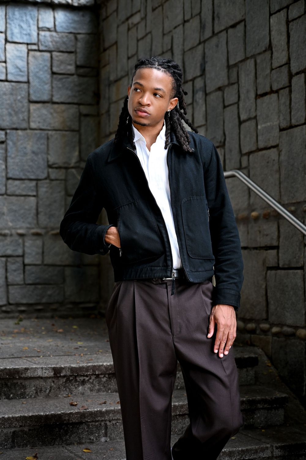 A person in a black jacket and brown pants poses against a stone wall in a series of four similar shots.