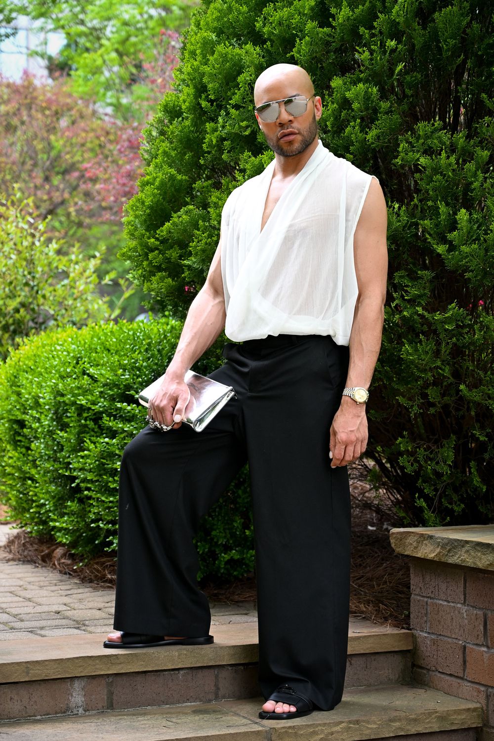 Person wearing a sleeveless white top and flowy black palazzo pants poses on outdoor stone steps near lush greenery.
