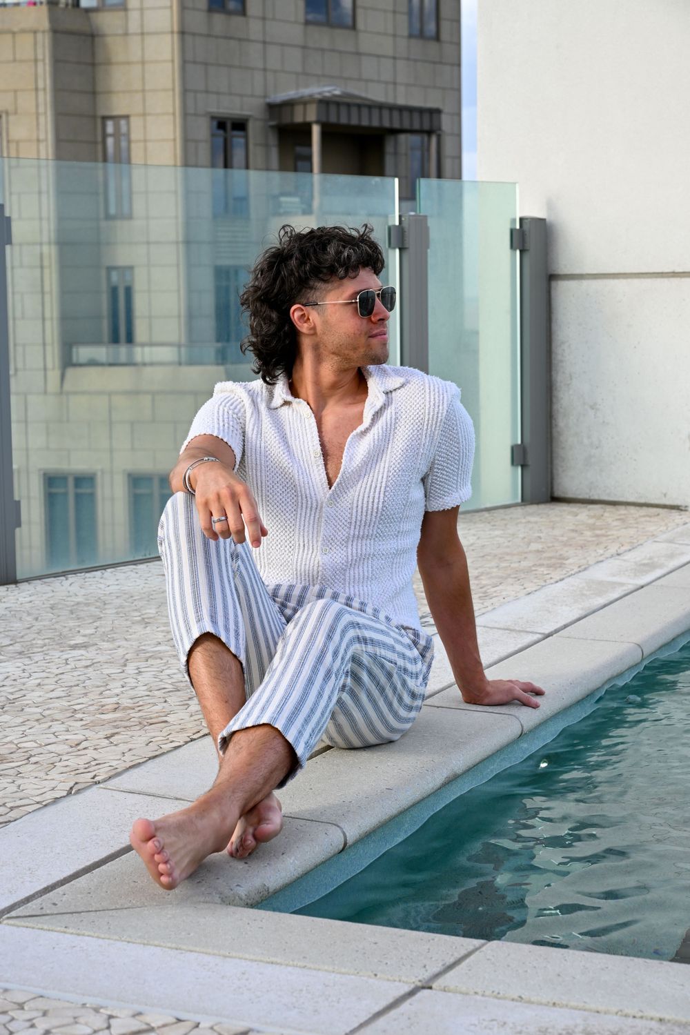 Stylish person in sunglasses and striped outfit relaxing by a rooftop pool on a sunny day.