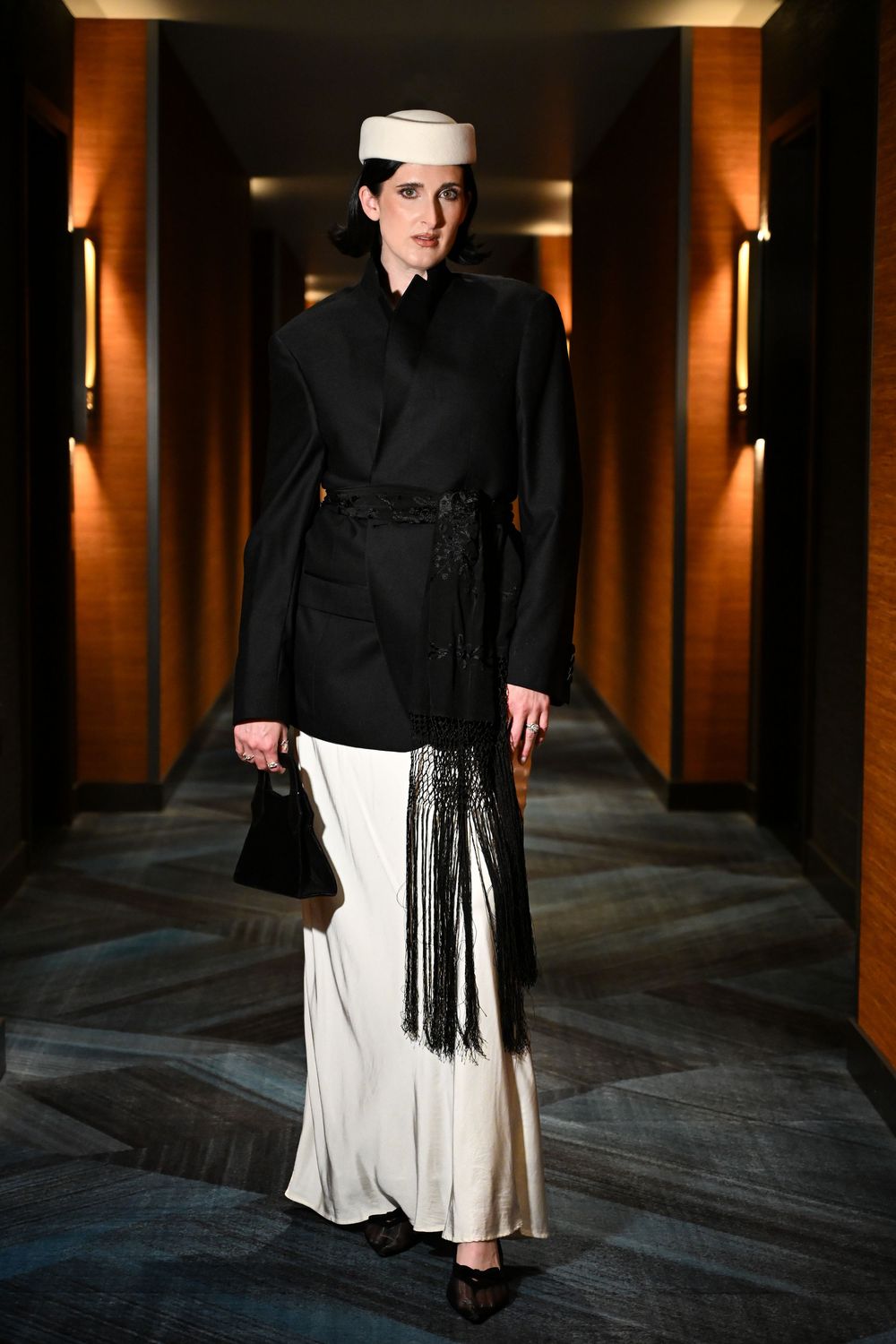 Model walks corridor in black belted blazer, white fringe skirt, white pillbox hat, carrying black handbag.