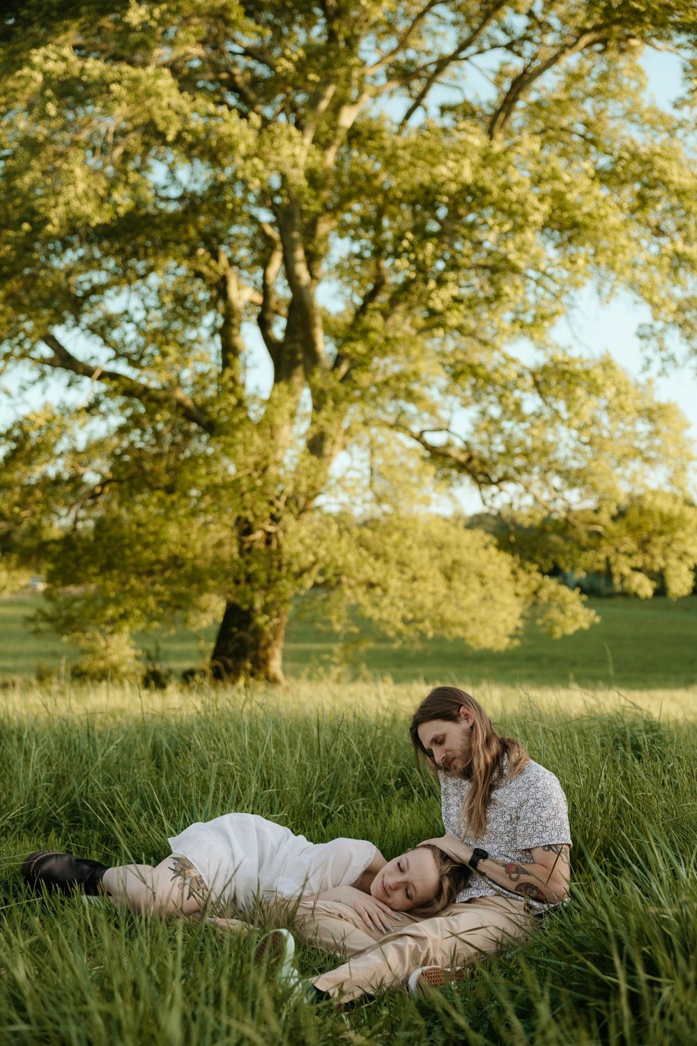Couple resting peacefully in tall grass beneath a large tree during golden hour.