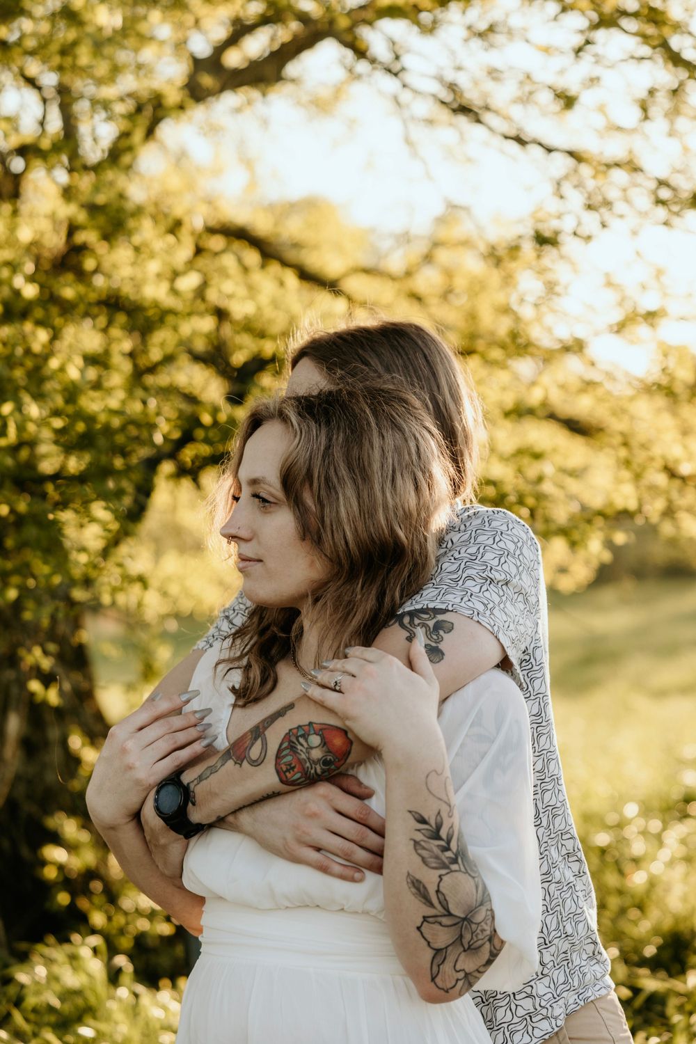 A tender embrace captured in golden sunlight with visible floral tattoos and casual attire.