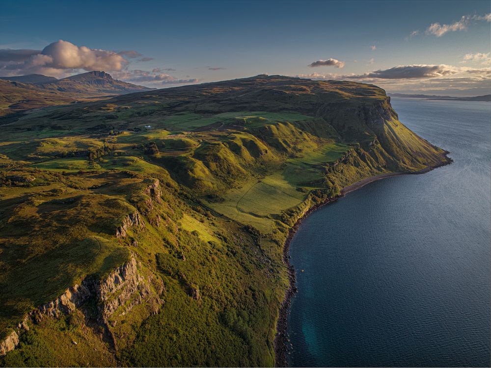 Dramatic coastal cliffs and rolling green hills overlooking deep blue waters during golden hour.