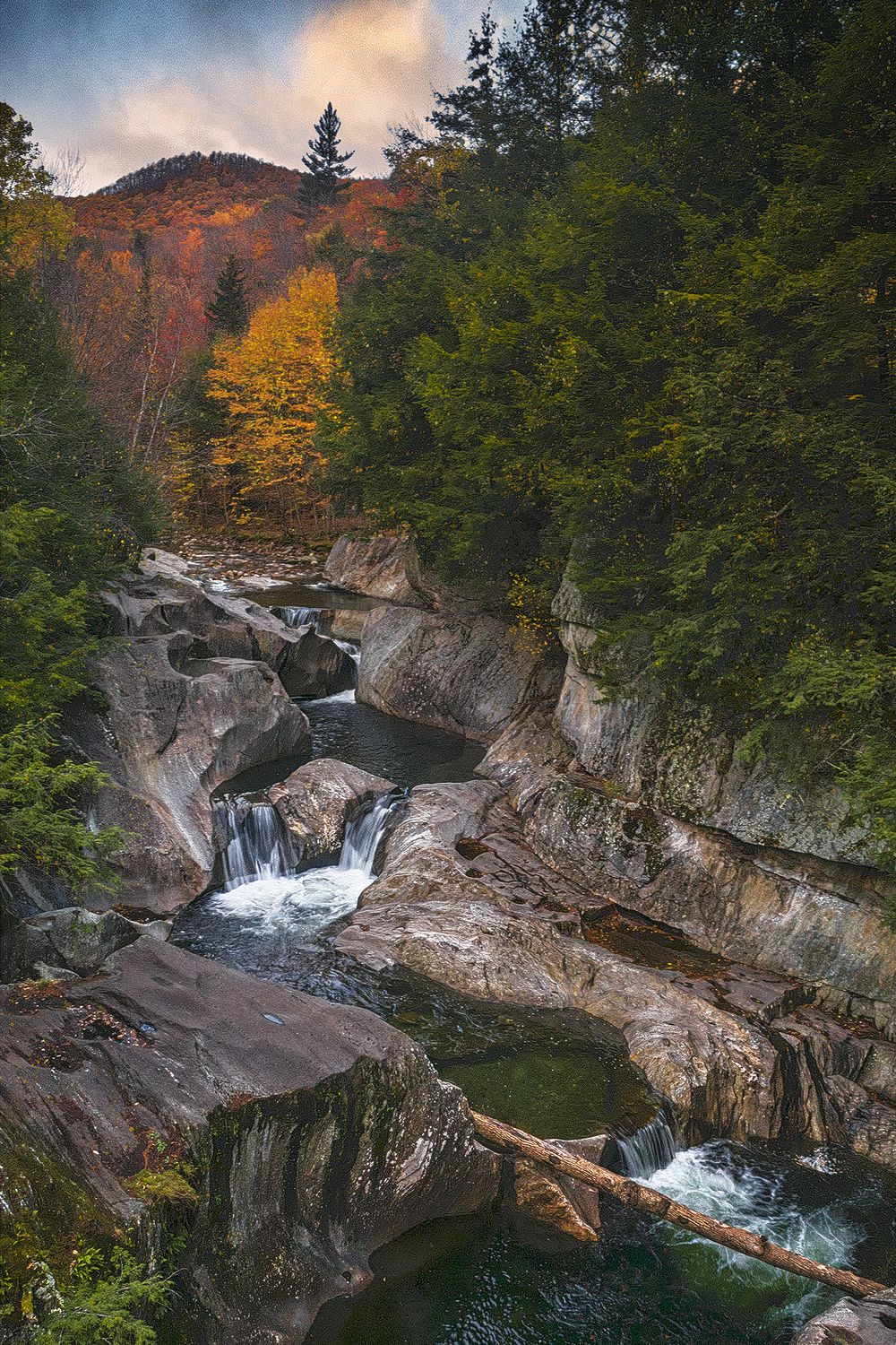 A scenic waterfall cascades through rocky gorge surrounded by autumn foliage and evergreen trees in a mountain landscape.