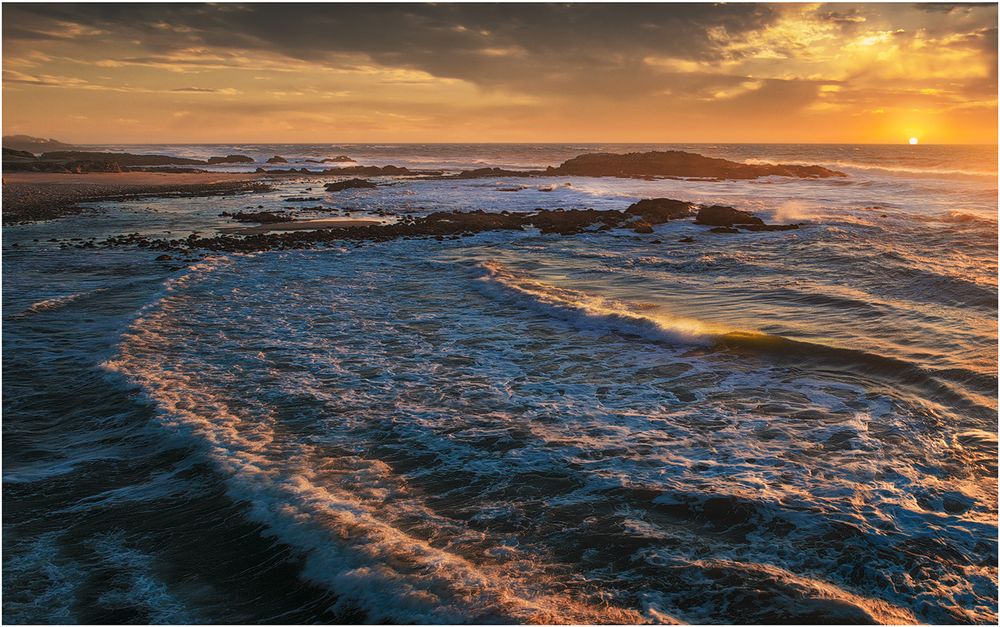 Dramatic ocean waves crash against rocky coastline during golden sunset with vibrant orange sky reflecting on water.