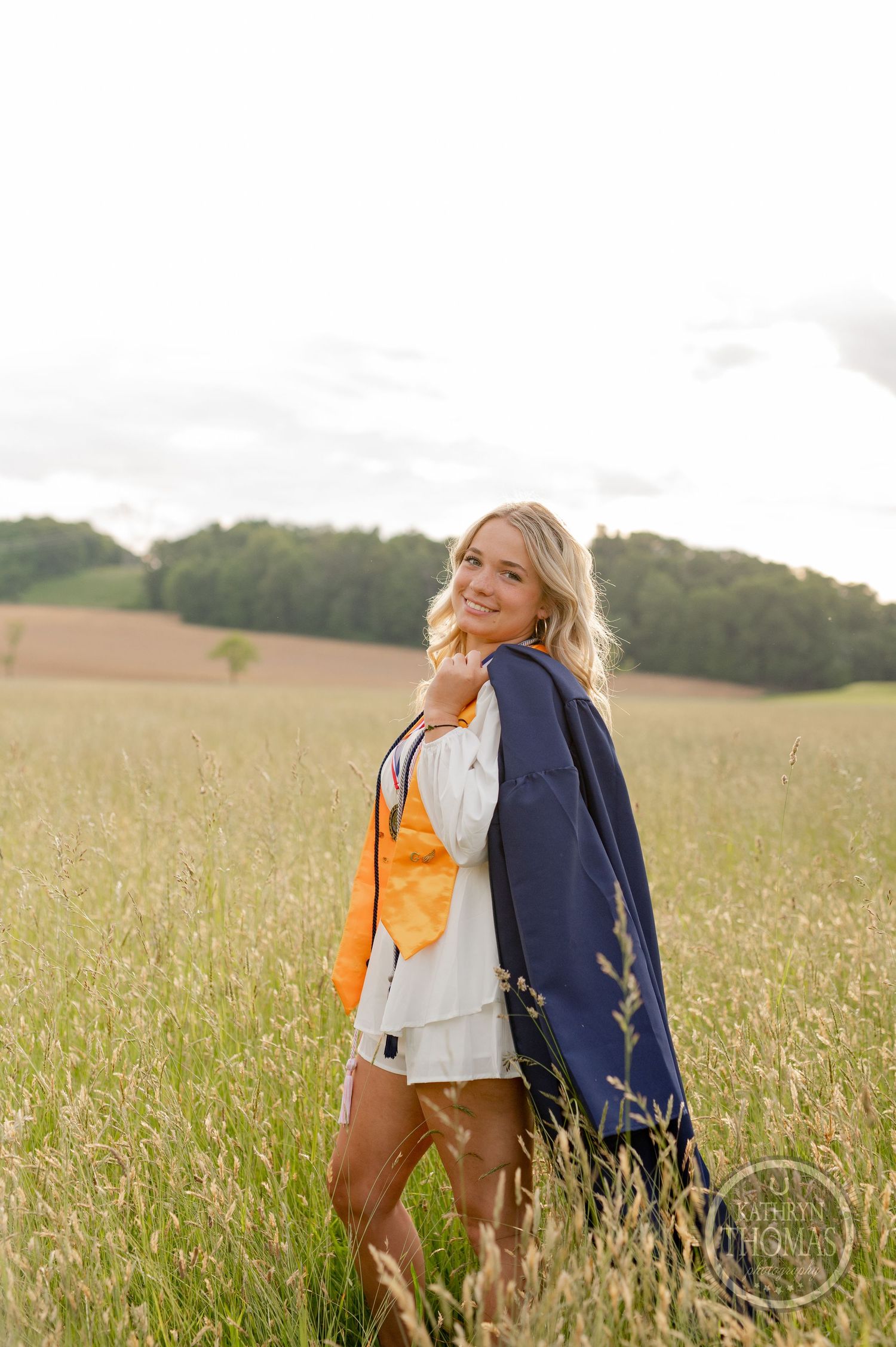 Graduate in academic dress poses in summer field during golden hour.