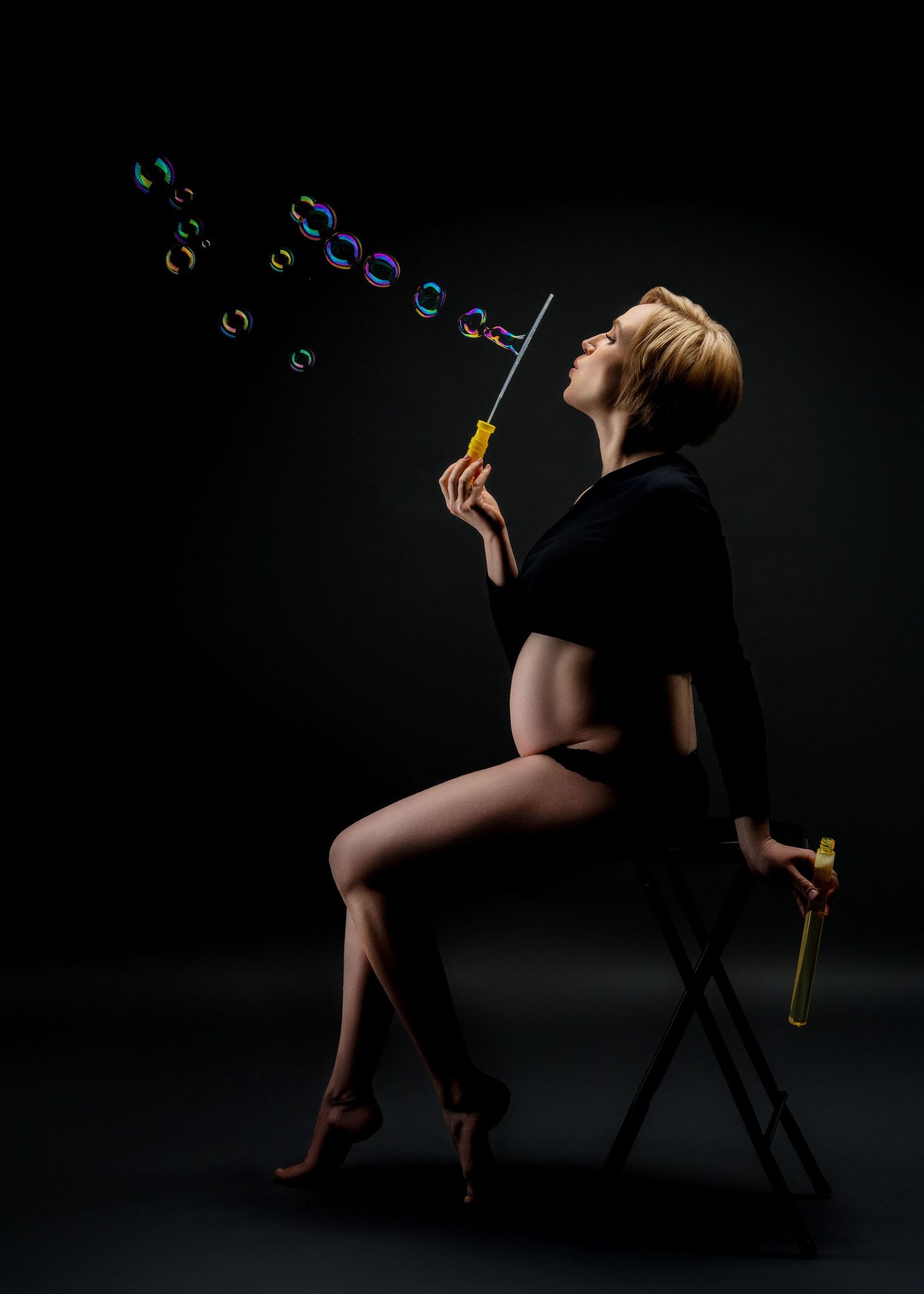 a pregnant mother sits in a dark studio with professional lighting and blows bubbles