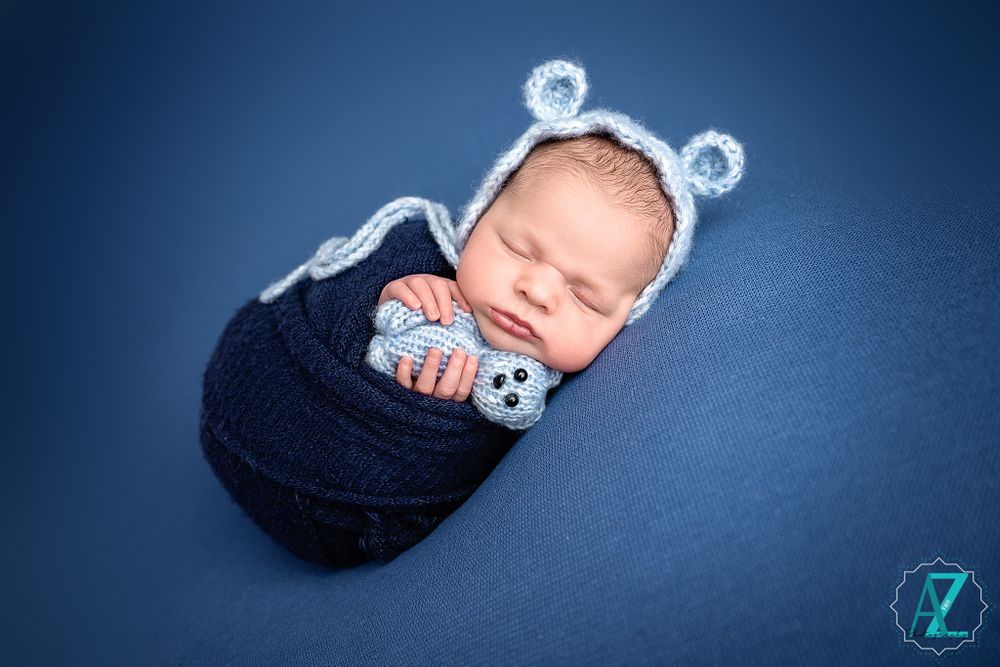 Newborn in bear ear hat and navy wrap sleeping on blue backdrop.